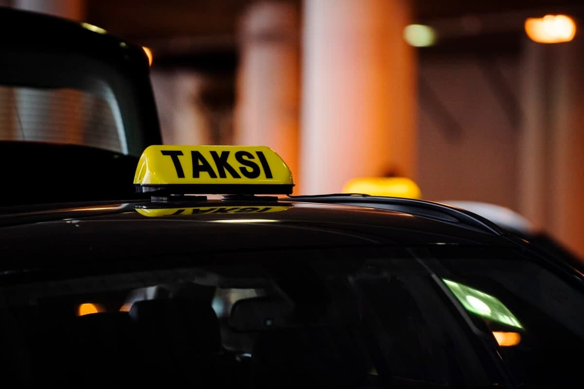 A lit yellow TAKSI roof sign on a dark taxi parked in a covered rank