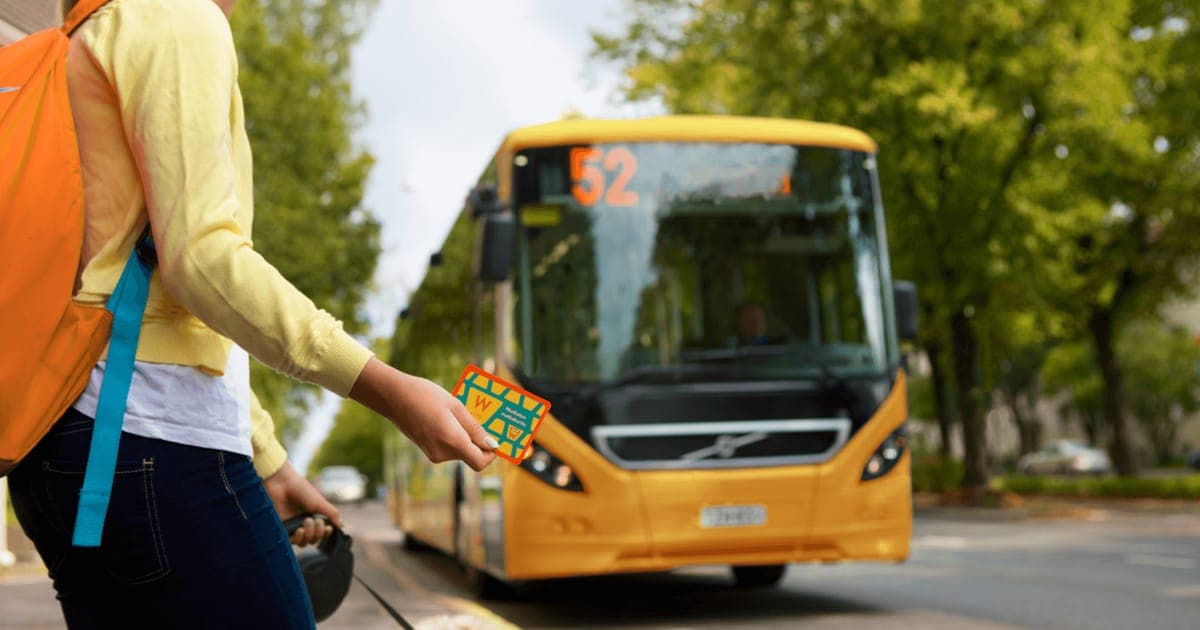 A passenger with an orange backpack holding out a Waltti travel card as a yellow number 52 bus pulls in