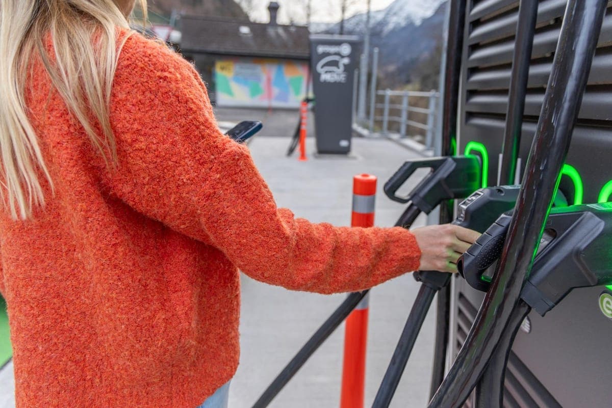 A person in an orange jumper lifting a charging connector from an EV fast charger at an outdoor station