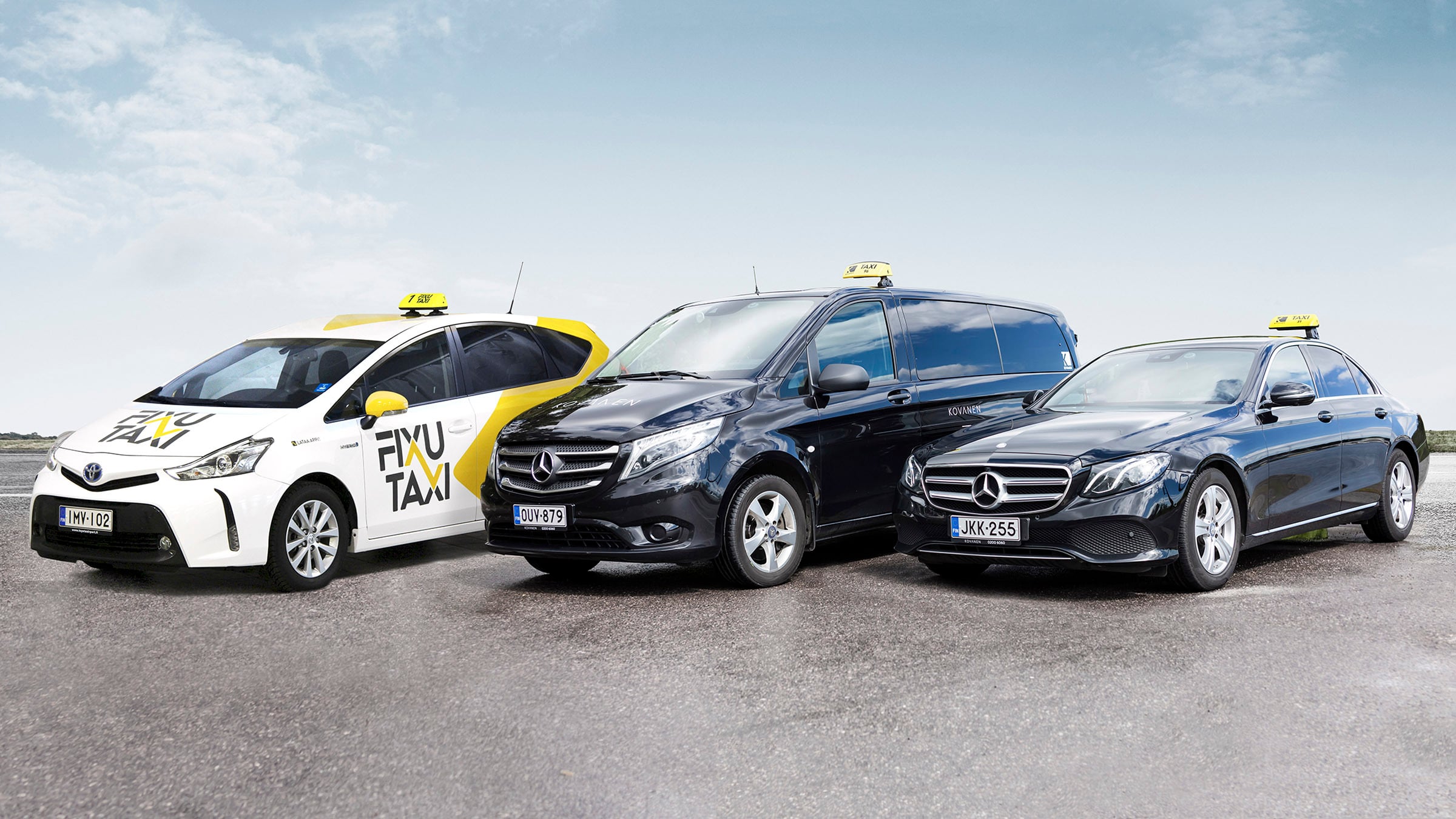A white and yellow FixuTaxi Toyota parked beside two black Kovanen Mercedes-Benz taxis, a van and a sedan, under a pale blue sky