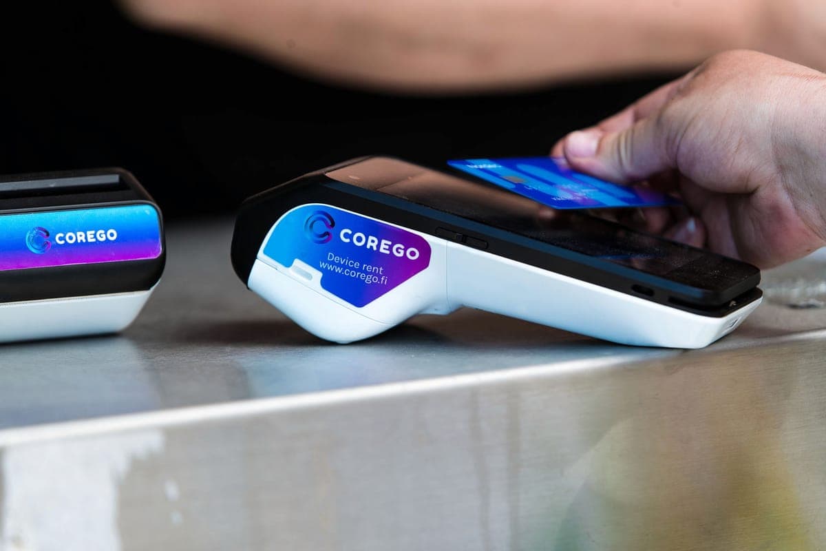 A hand tapping a bank card against a Corego-branded payment terminal on a metal counter