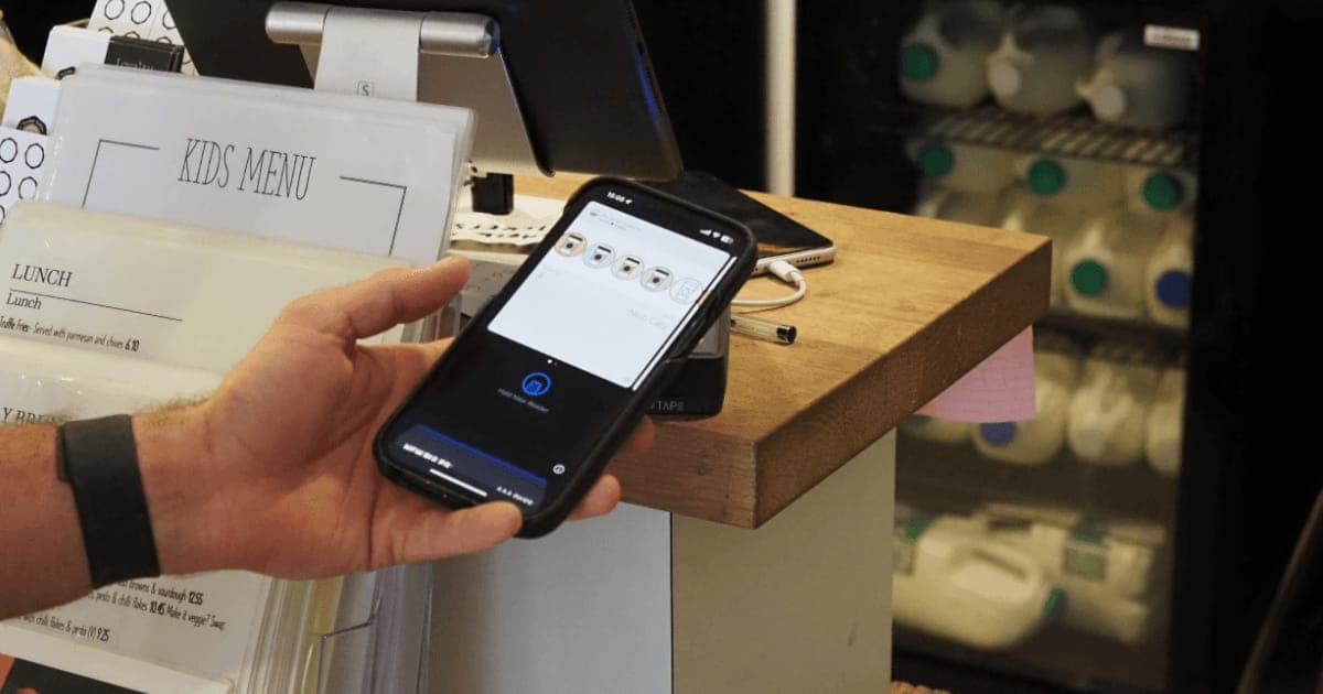 A hand holding a phone above an NFC reader on a cafe counter, beside printed lunch and kids menus