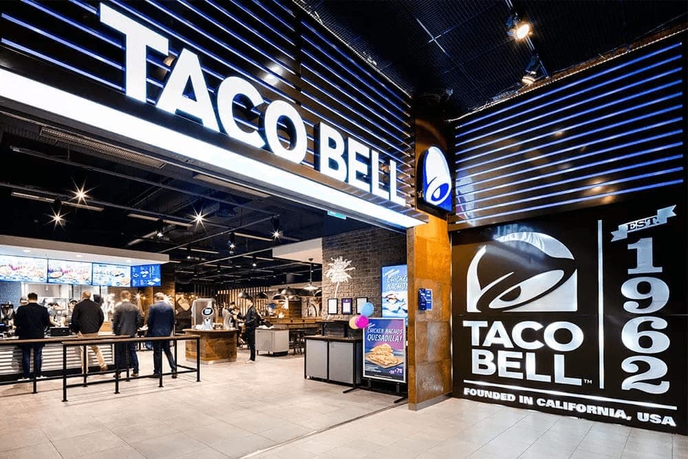 The interior of a Taco Bell restaurant, customers queueing at the counter under an illuminated brand sign
