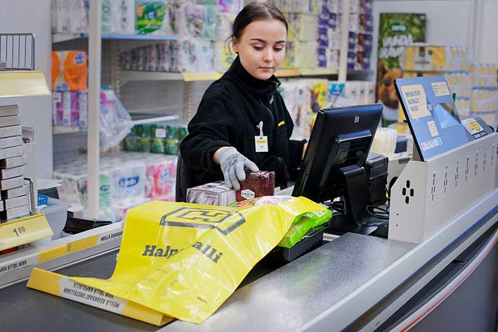 A Halpa-Halli cashier packing shopping into a yellow branded carrier bag at the checkout