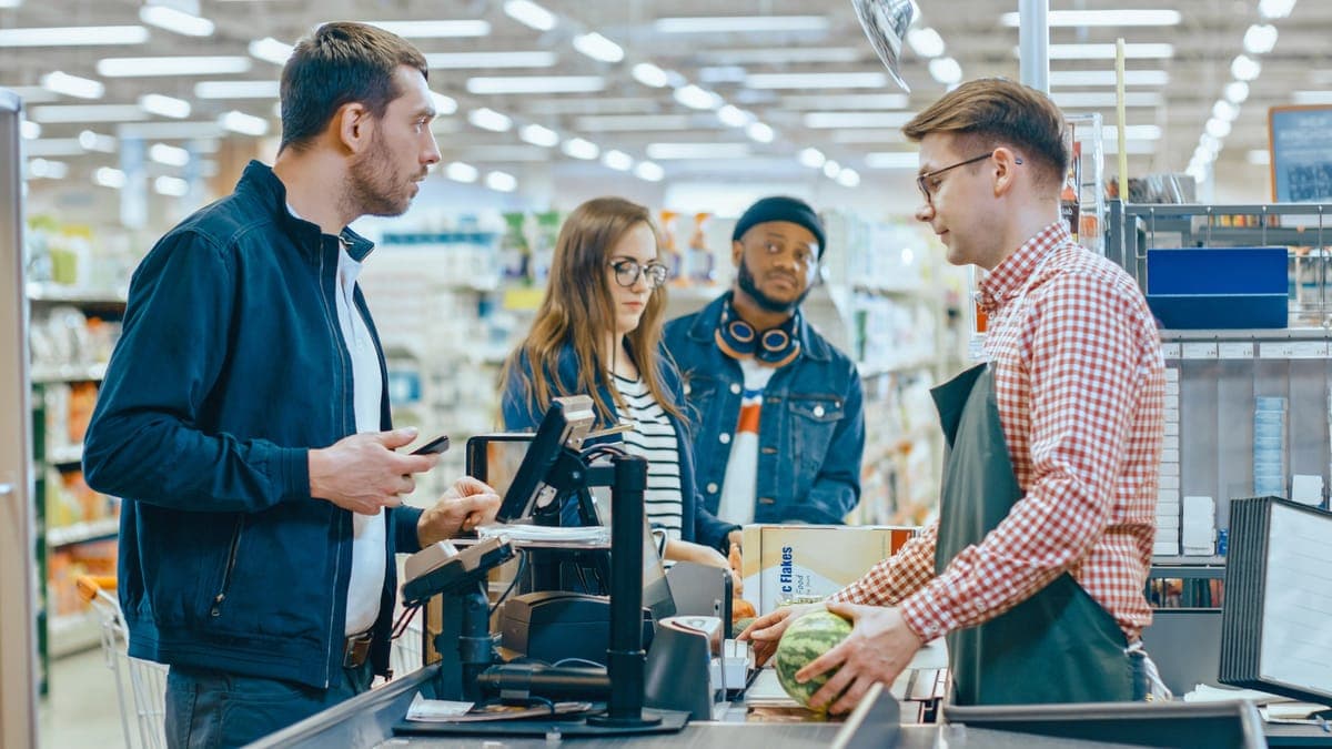 A customer paying by phone at a supermarket checkout while the cashier scans a watermelon