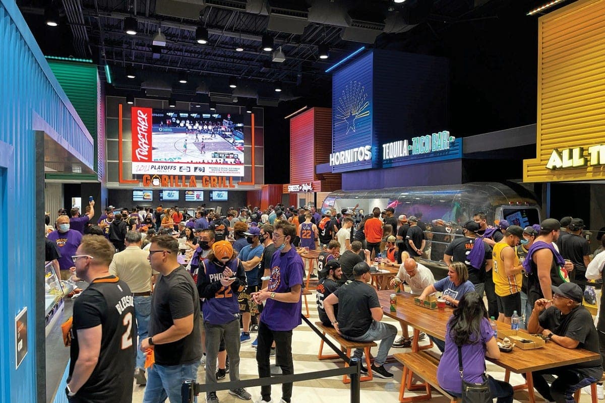 A crowded stadium concourse food hall, fans in team shirts queueing at counters below a screen showing the game