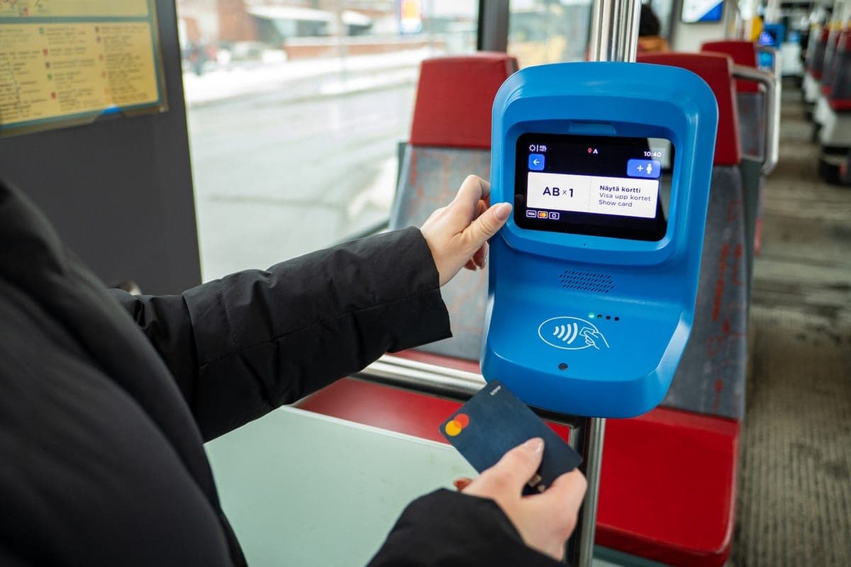 A passenger holding a bank card up to a blue HSL card reader on a tram, the screen reading AB times 1