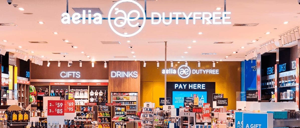 The interior of an Aelia Duty Free store, with lit brand signage above gift, drinks and pay here counters