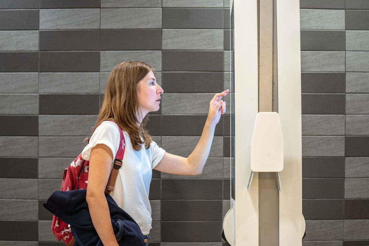 A shopper with a red backpack reaching up to tap a wall-mounted touchscreen in a store