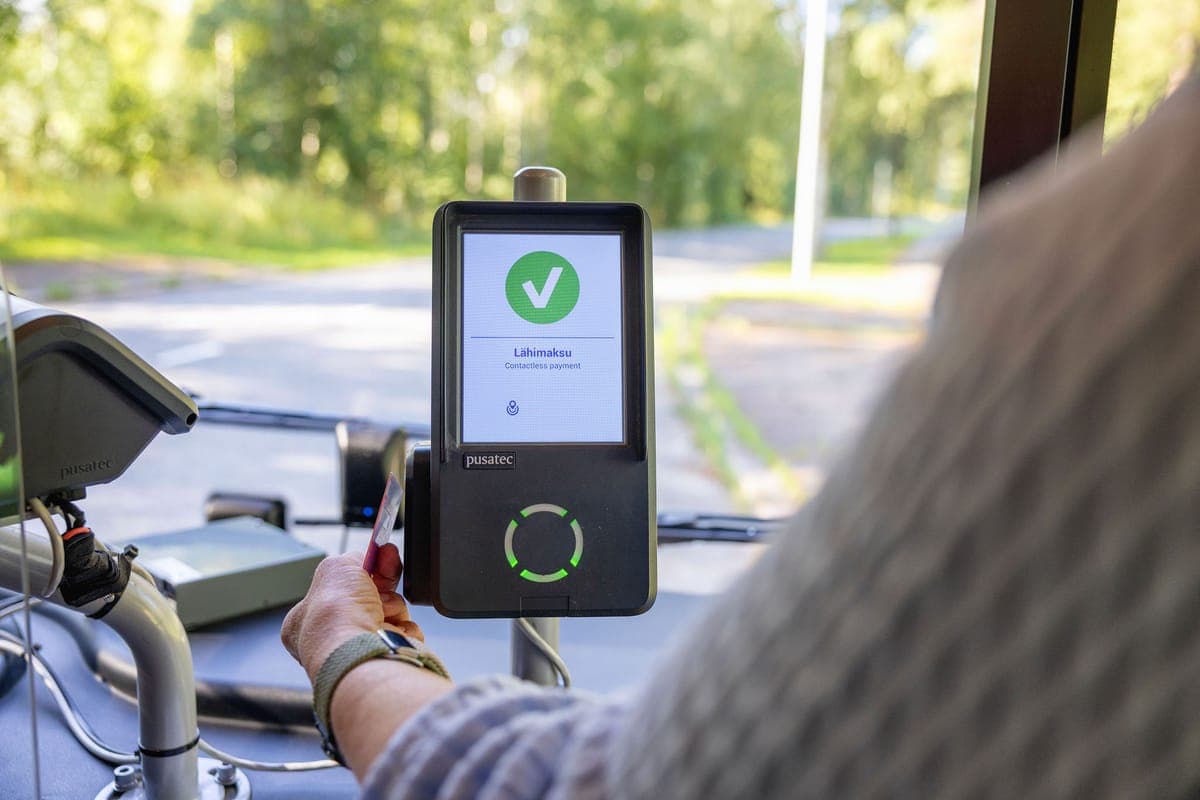 A passenger tapping a card on a Pusatec bus reader, the screen showing a green tick and the word Lahimaksu, contactless payment