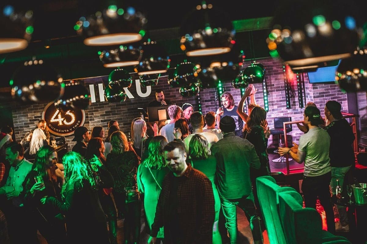 A busy bar under green lighting and mirror balls, with people drinking and a band on the stage behind