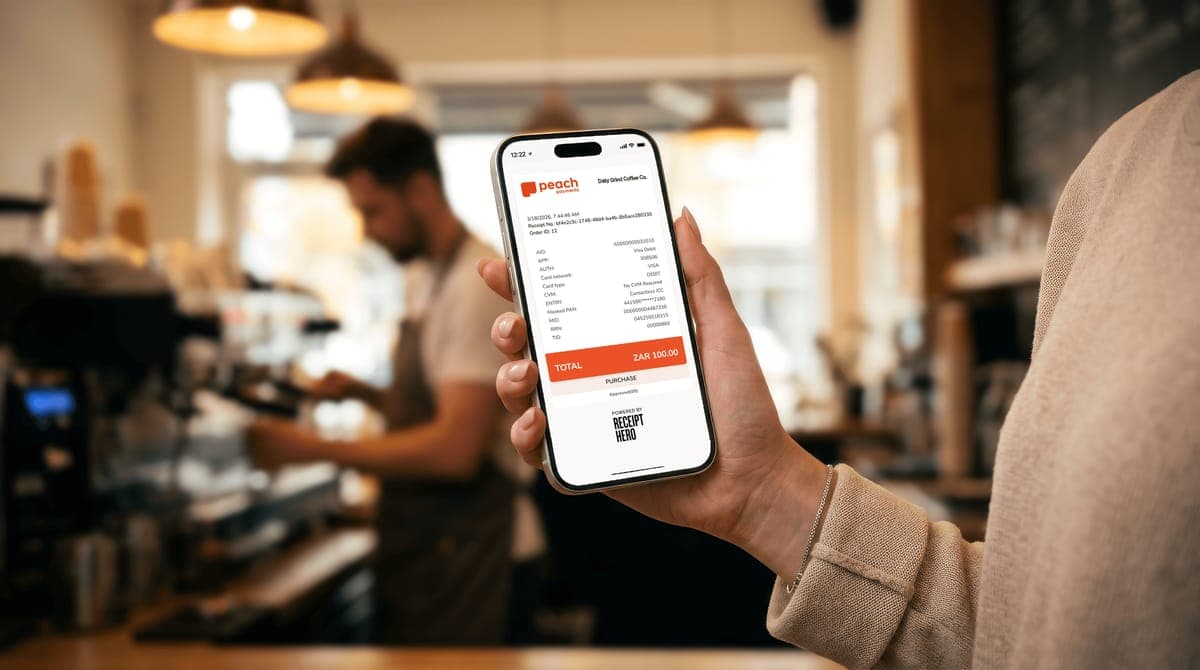 A hand holding a phone showing a Peach Payments digital receipt for a coffee shop, total ZAR 100, badged Powered by ReceiptHero, with a barista behind