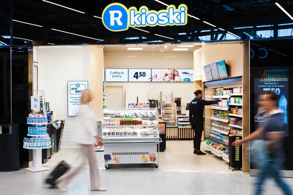 An R-kioski in a station concourse, a member of staff stocking shelves while travellers pass by