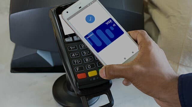 A hand holding a phone showing a Nordea Visa card in a mobile wallet against a card terminal