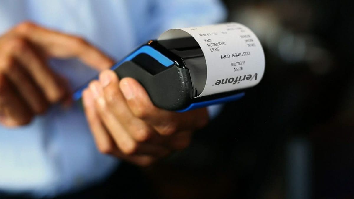 A hand holding a Verifone card terminal as a paper customer copy receipt prints from it