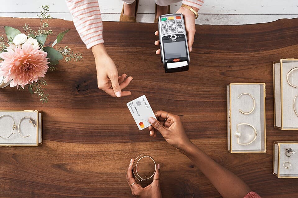 Seen from above, a customer handing a Mastercard to a jeweller holding a card terminal across a wooden counter