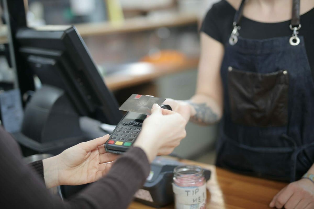 A customer tapping a card on a terminal held by a cafe worker in an apron, a tip jar on the counter