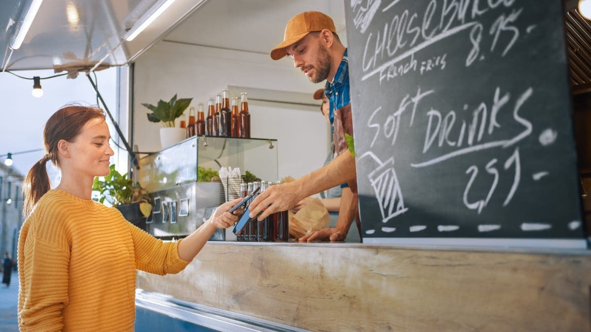 A customer in a yellow jumper paying by phone at a food truck counter, the menu chalked on a board beside her