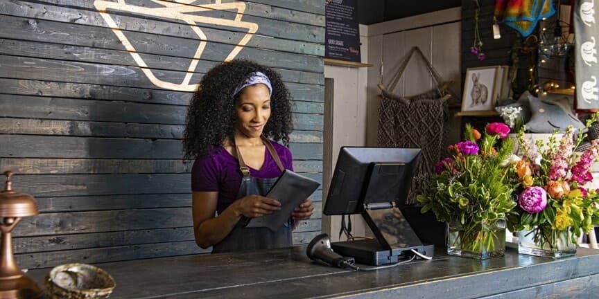 A shop assistant at the counter of a florist, holding a tablet beside a till, with bouquets on the counter