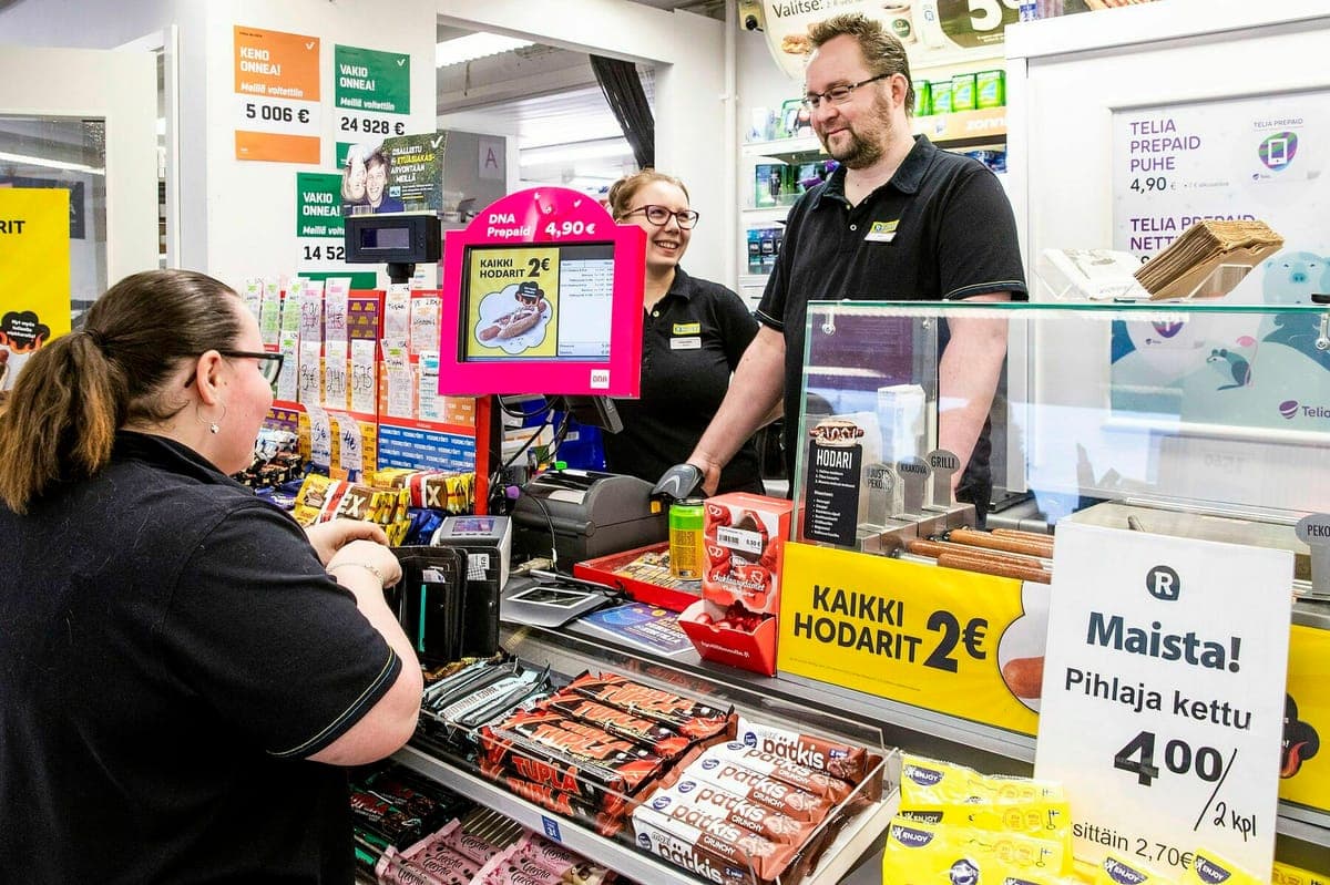 A customer at an R-kioski counter being served by two staff, hot dog and prepaid offers displayed around the till