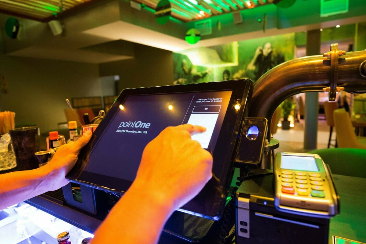 A bartender logging into a pointOne till screen behind a green-lit bar, a card terminal beside it