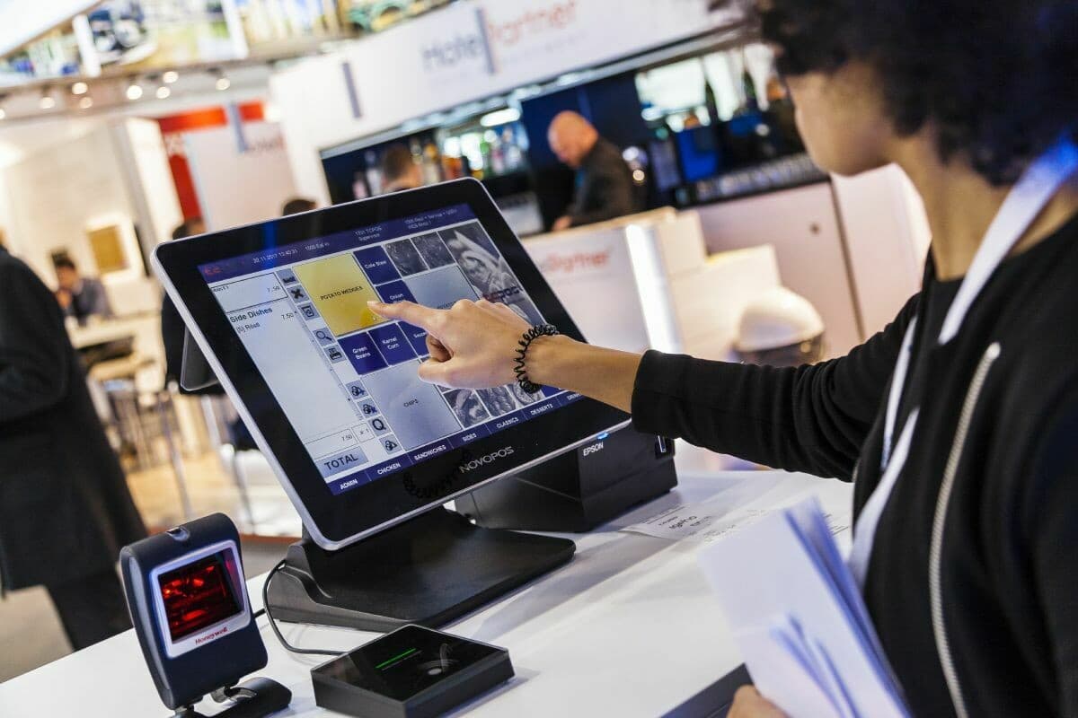 A visitor at a trade stand tapping a menu of side dishes on a NovoPOS touchscreen till, a barcode scanner in front
