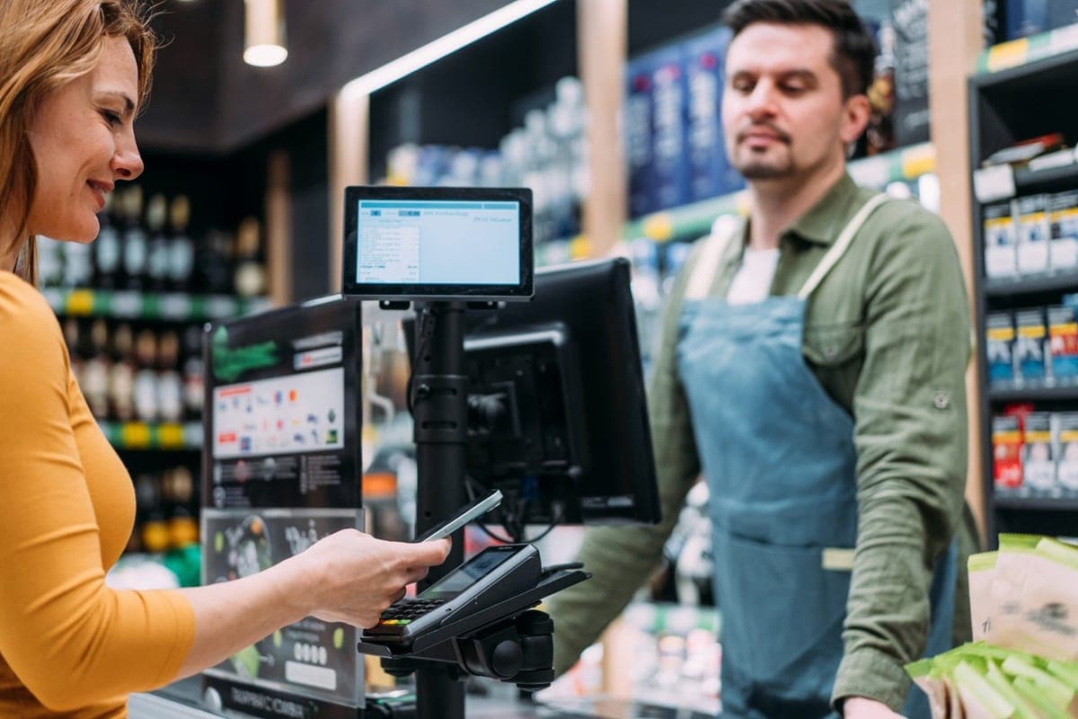 A shopper paying by phone at a grocery counter while the assistant looks on
