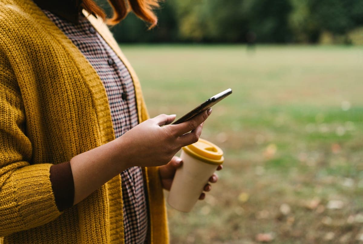 A woman in a mustard cardigan holding a phone and a reusable coffee cup in a park