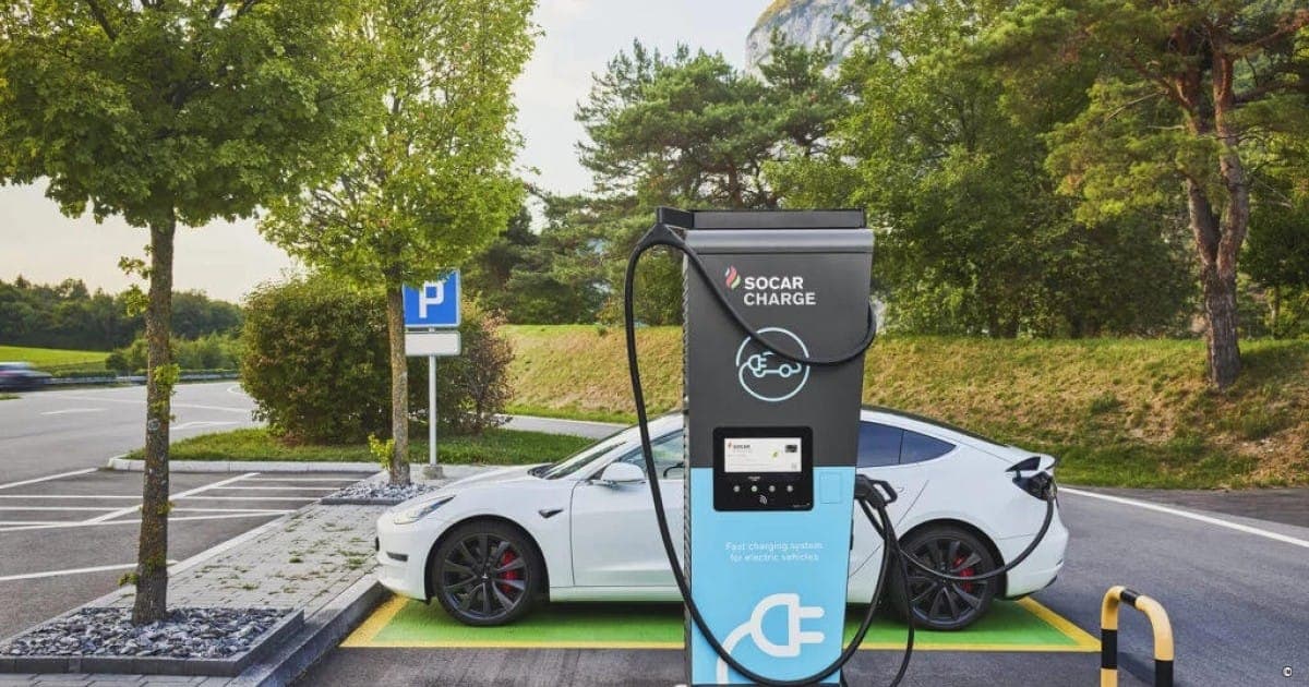 A white electric car plugged into a Socar Charge fast charger in a tree-lined car park