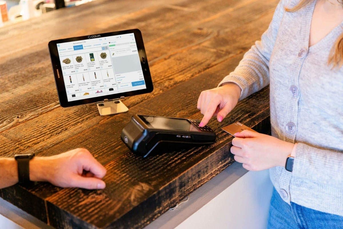 A customer holding a card beside a terminal on a wooden counter while the assistant enters the amount, a Cova tablet till alongside