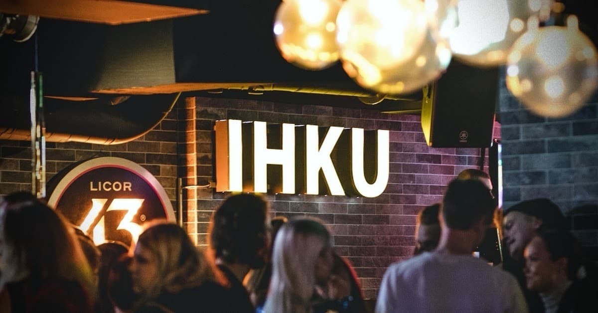 A crowded bar under mirror-ball lights, with an illuminated IHKU sign on the brick wall behind