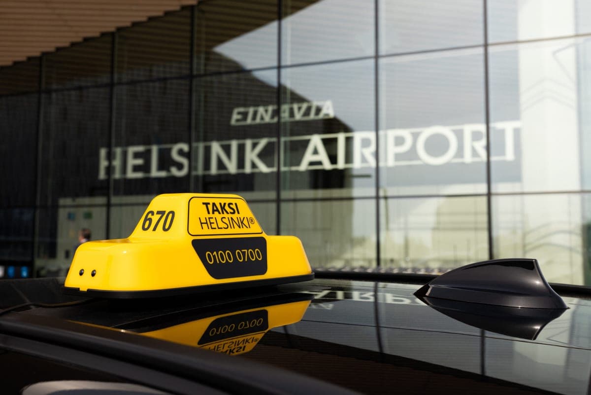 A yellow Taksi Helsinki roof sign on a car outside the glass frontage of Helsinki Airport