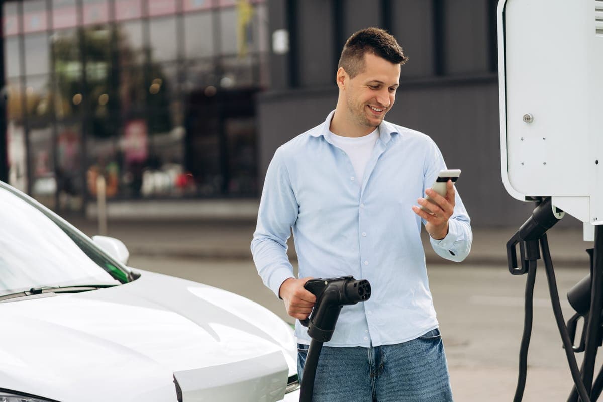 A driver at a public charging station holding a charging connector in one hand and his phone in the other, beside a white electric car
