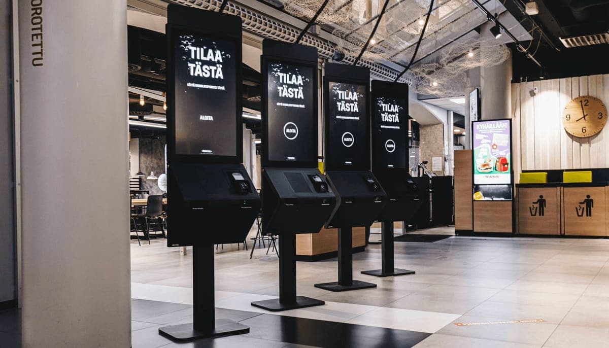A row of four self-order kiosks in a quick-service restaurant, each screen reading Tilaa tasta
