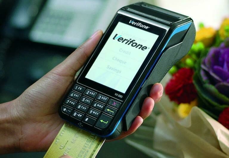A hand holding a Verifone VX 690 card terminal with a card inserted, a bunch of flowers behind