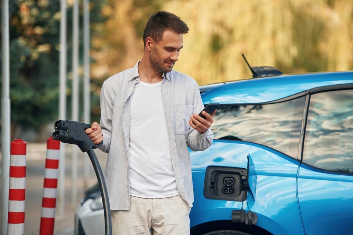 A man holding a charging connector and looking at his phone beside a blue electric car