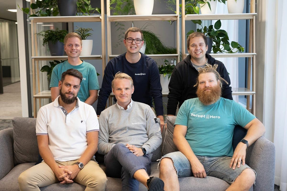 Six members of the ReceiptHero team in branded shirts, seated and standing in front of a plant-filled shelving unit