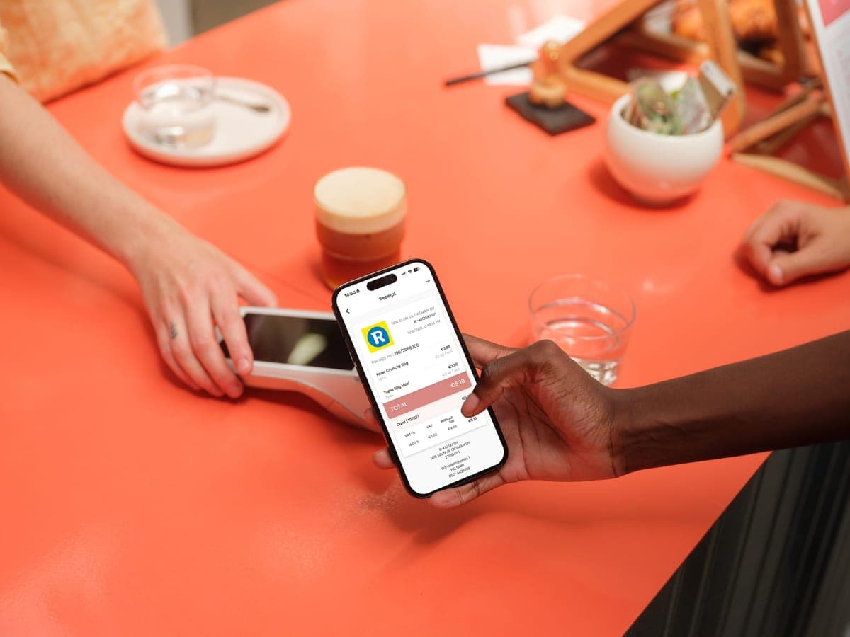 A customer holding a phone showing an R-kioski digital receipt while paying by card at a red counter