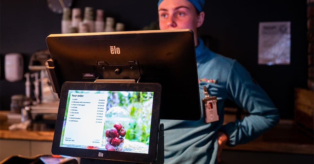 A cafe worker behind an Elo touchscreen till, the customer-facing screen listing the order